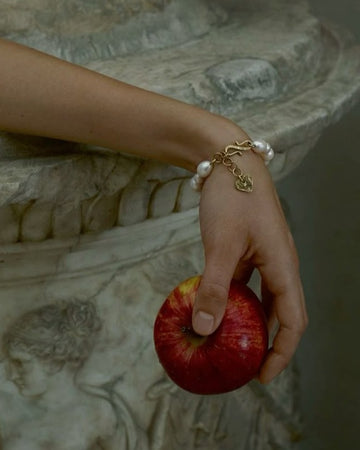 Hand holding a red apple with a pearl bracelet against an ornate stone background