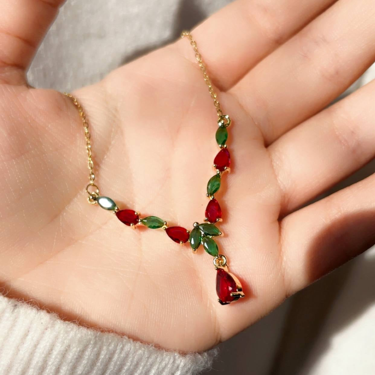 Gold necklace with red and green gemstones held in a hand against a neutral background
