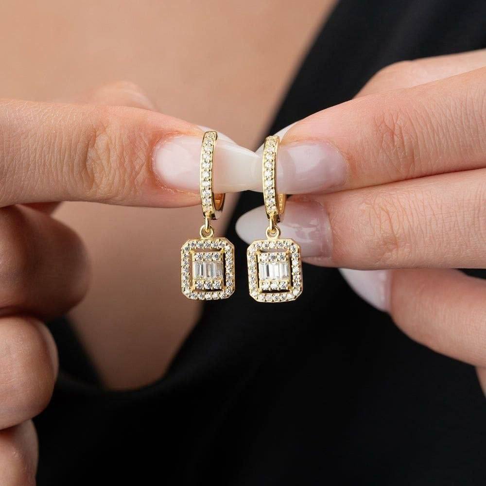 Gold earrings with square-cut gemstones held between fingers against a neutral background